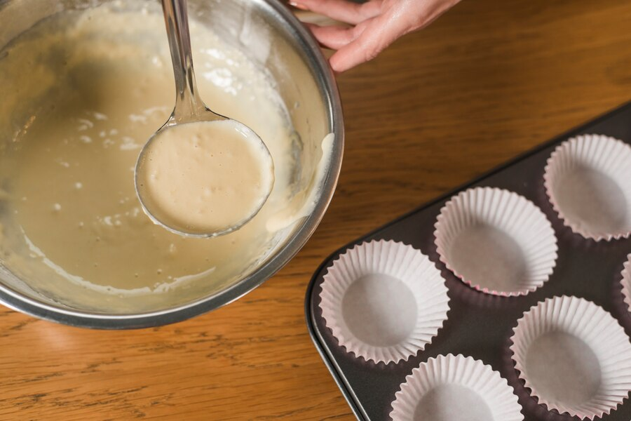 Scooping cake batter from a bowl and a cupcake pan with cupcake liners placed on it