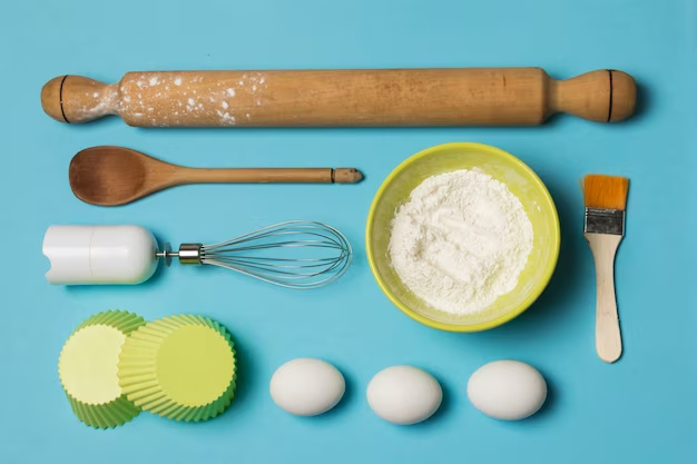 rolling pin, whisk, wooden ladle, brush, parchment paper, flour, and eggs on a table